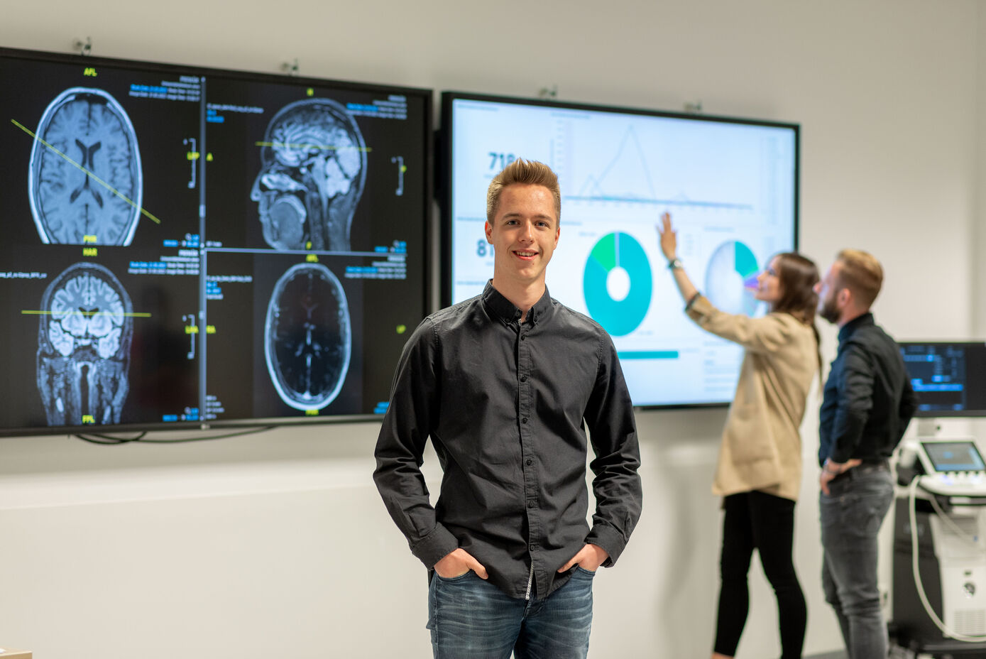 Symbolbild: Ausbildung zum Fachinformatiker oder Fachinformatikerin für Systemintegration - männlich gelesene Person steht im Vordergrund der das Bild betrachtenden Person zugewandt. Im Hintergrund stehen verschwommen zwei Personen die sich Inhalte auf einem großen Bildschirm anschauen.