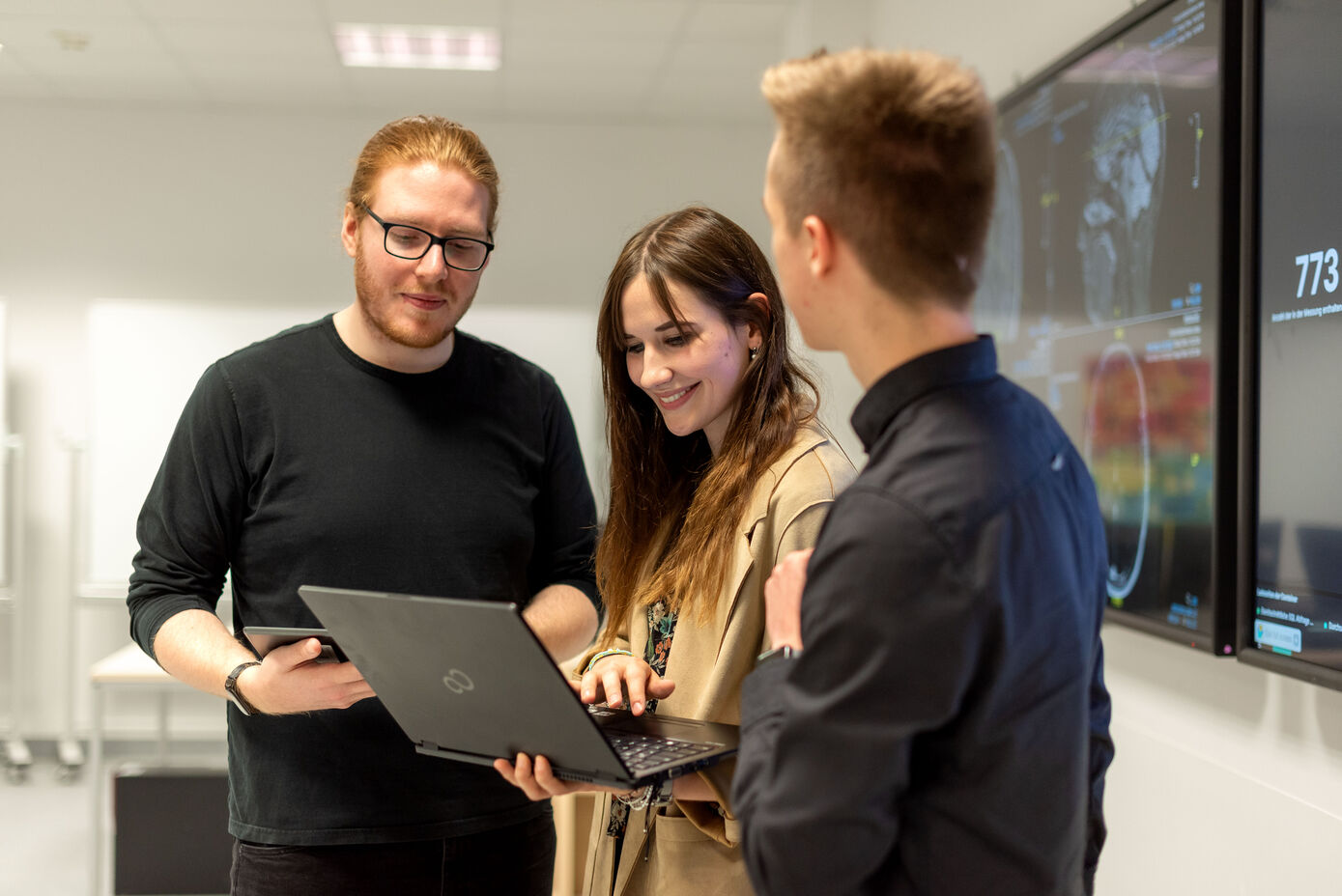 Symbolbild: Ausbildung zum Informatikkaufmann oder zur Informatikkauffrau - drei junge Personen stehen zusammen und schauen auf einen Laptop, den eine Person von ihnen in den Händen hält.