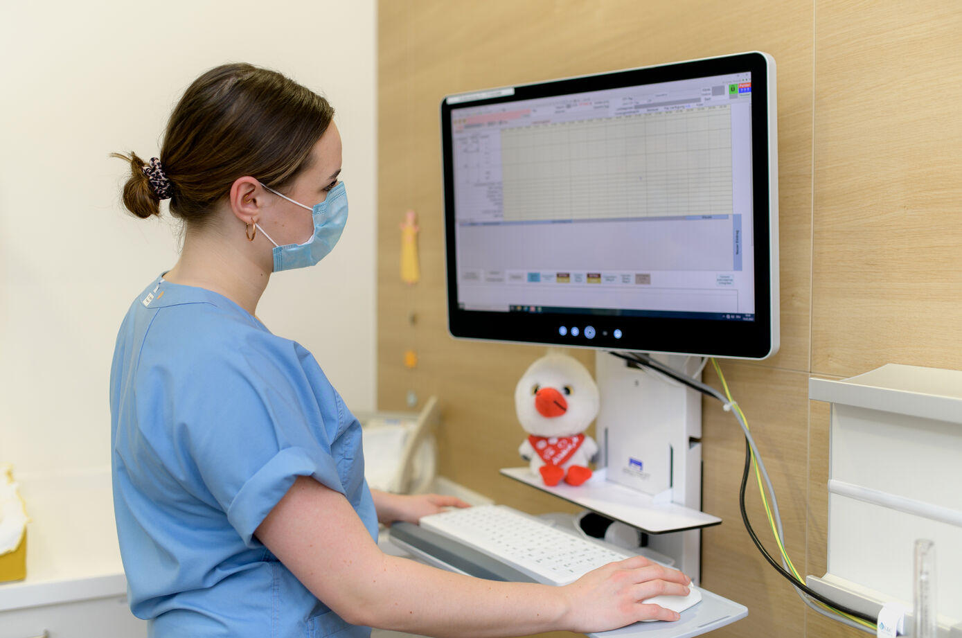 Symbolbild für das duale Studium zur Hebamme: eine Hebamme steht an einem PC und dokumentiert die Wehen einer Patientin in der Patientenakte.