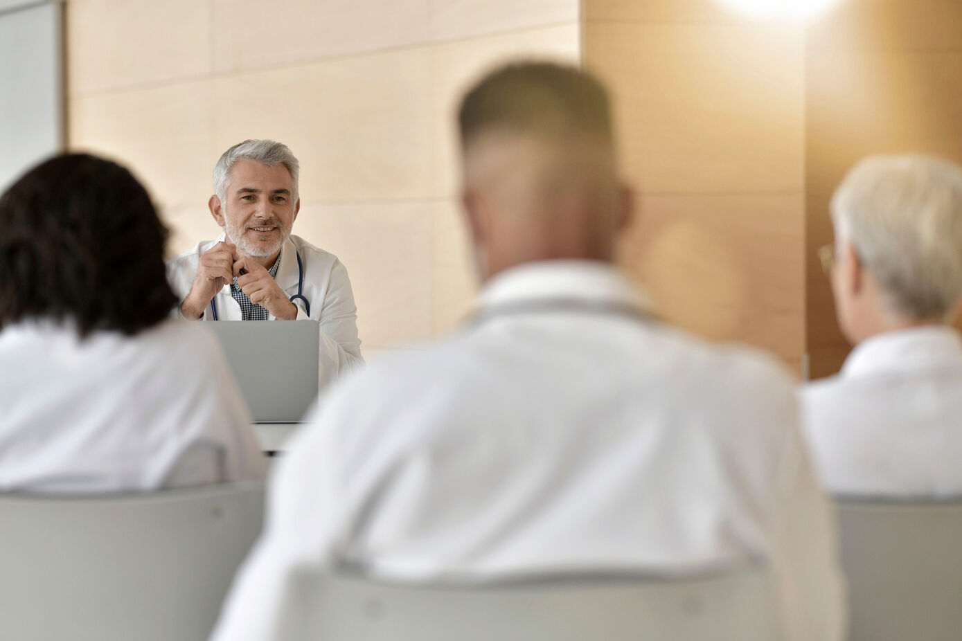 Symbolbild für Stellenausschreibungen von Professuren der Medizinischen Fakultät: Seminarsituation, eine männlich gelesene Person fortgeschrittenen Alters (graue Haare) sitzt in einem Raum vorn, er hat einen weißen Kittel an, sowie ein Stethoskop um den Hals. Ihm zugewandt sitzen drei Personen mit dem Rücken zur Person, die dieses Bild betrachtet. Auch sie haben weiße Kittel an.