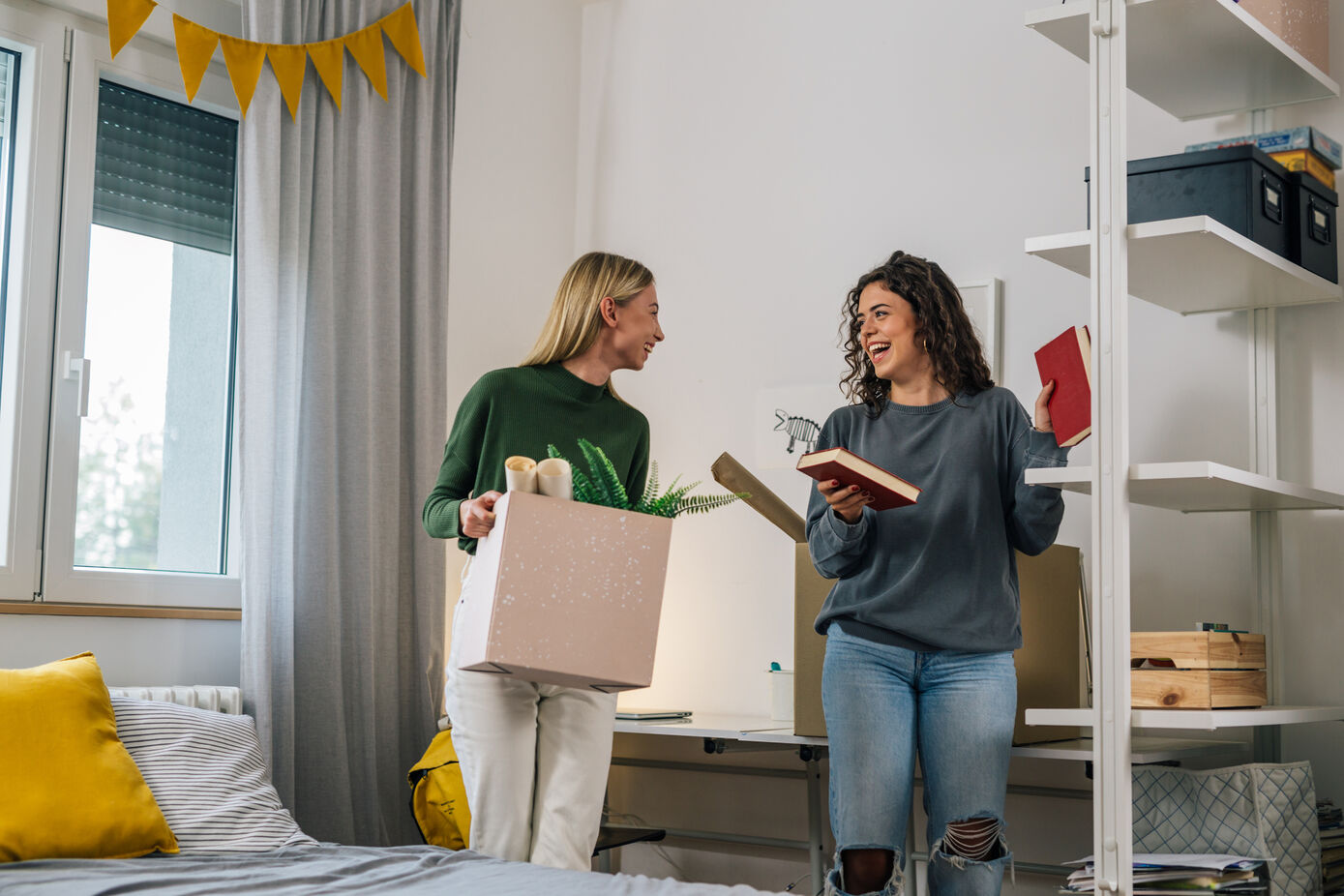 Symbolbild für Umzug: zwei weiblich gelesene Personen stehen in einem Zimmer und unterhalten sich, die links stehende Person hält einen Umzugskarton in der Hand, die rechts stehende Person stellt Bücher in ein Regal.